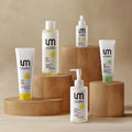 Five UM Lesimo skincare bottles and tubes arranged on wooden display blocks against a warm beige background. The products include cleansers, toners, and serums in white packaging with yellow and green accents.